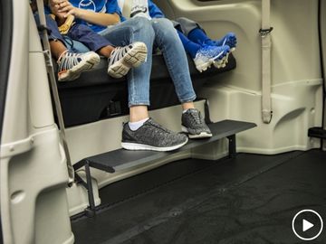 Three people seated in thrid-row of BraunAbility Chrysler Pacifica wheelchair van utilizing the foot rest feature. Play-video symbol in lower left corner.