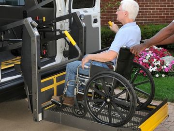 Man using manual wheelchair is being assisted onto a platform wheelchair lift to enter a full-size van