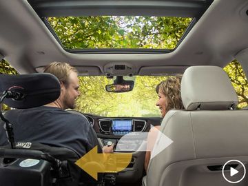 Driver and front passenger in a VMI Northstar Chrysler Pacifica wheelchair van. The driver is in a power wheelchair. Overlay graphic of double arrows to deomontrate seats are interchangeable.