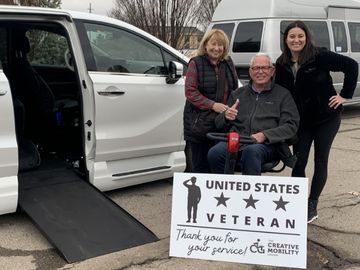 Disabled veteran using a mobility scooter with two other people in front of a wheelchair accessible minivan with the ramp deployed and holding a sign that reads "United States Veteran. Thank you for your service!" with The Creative Mobility Group logo.