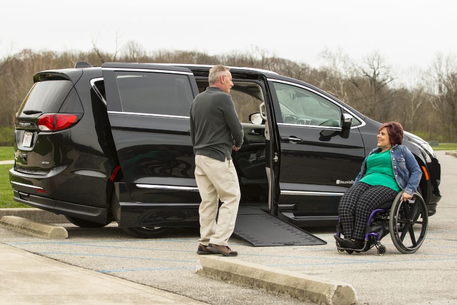Man and woman using a manual wheelchair in a parking lot beside a wheelchair accessible Chrysler Pacifica with a wheelchair ramp deployed from the passenger-side sliding door.