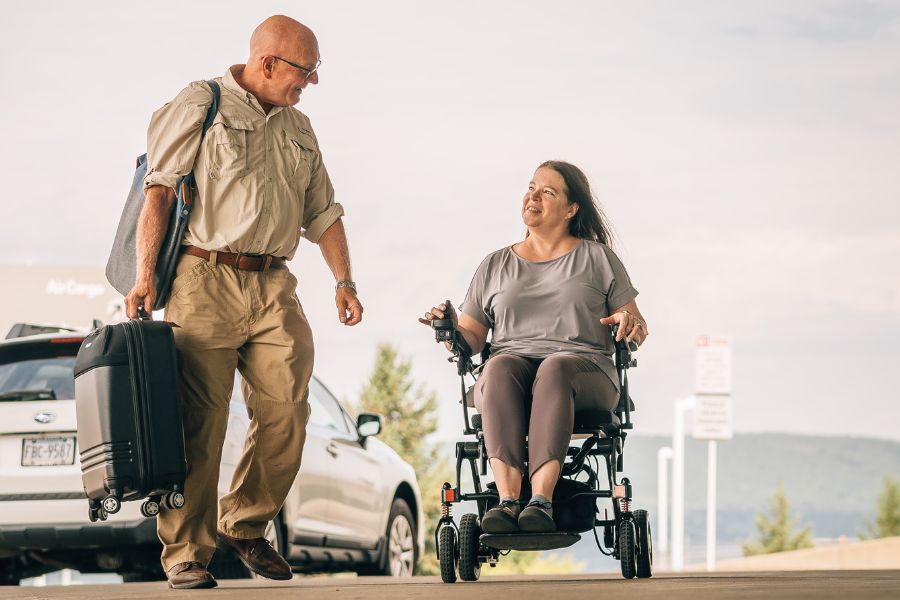 Woman operates power wheelchair while a man carrying luggage walks beside her