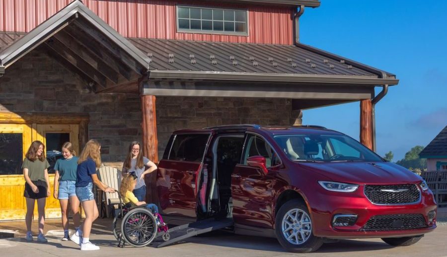 Child using a manual wheelchair is assisted up a ramp into a side-entry wheelchair accessible van