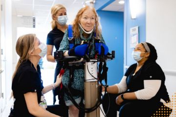 Three healthcare workers assist a patient on a piece of rehabilitation equipment.