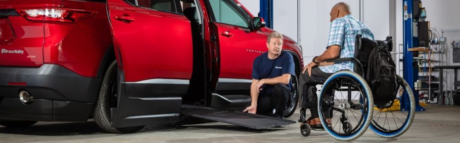 Service technician kneeling beside a wheelchair accessible BraunAbility Chevrolet Traverse with ramp deployed, conversing with customer seated in wheelchair.