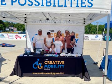 A group of people stands behind a booth for The Creative Mobility Group at an outdoor event. The booth features a table with promotional materials and a banner with the company's logo and website.