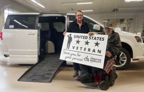 Veteran in a power wheelchair receives a wheelchair accessible van from The Creative Mobility Group with ramp deployed and thank you sign for military service