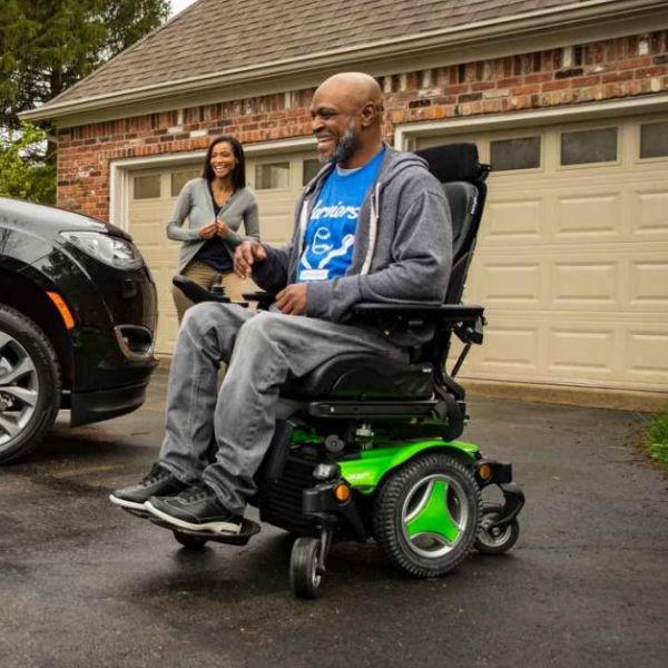 Man using a power wheelchair near an accessible vehicle at home with caregiver support, representing mobility solutions for loved ones